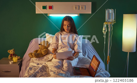 Teen girl in home wear sitting in bed in hospital ward and drawing with colorful crayons placed in wooden box, in modern clinic with medical equipment Teen girl in home wear sitting in bed in hospital ward and drawing with colorful crayons placed in wooden box, in modern clinic with medical equipment 122403186