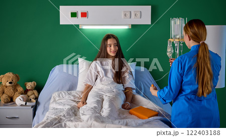 Teen girl sitting in bed in hospital ward while female nurse in blue medical gown doing IV drip procedure in modern clinic. Taking care after patient Teen girl sitting in bed in hospital ward while female nurse in blue medical gown doing IV drip procedure in modern clinic. Taking care after patient 122403188