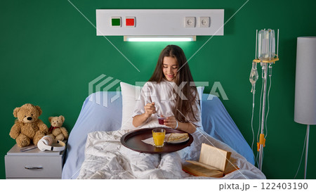 Teen girl sitting in bed in hospital clinic, with food tray and book and eating jelly for dinner. Modern well-equipped clinic and comfort service for patient 122403190