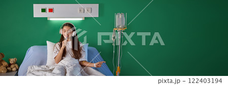 Smiling teen girl sitting in bed with white sheets, in comfortable hospital ward, listening to music in headphones and singing against green wall 122403194