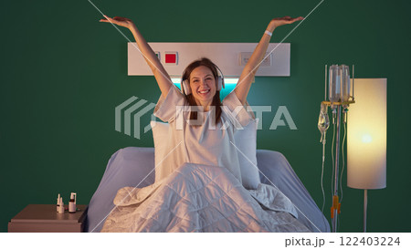 Happy, smiling young woman in medical gown sitting in bed in modern hospital ward, in headphones, raising hands upwards in happiness of recovery 122403224