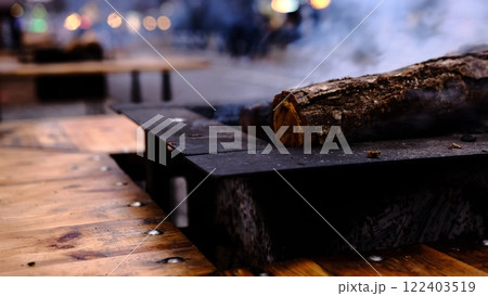 A glowing log burns gently in a stylish fire pit, creating a warm and festive atmosphere at a Christmas market while providing cozy heat for visitors on a chilly winter evening. A glowing log burns gently in a stylish fire pit, creating a warm and festive atmosphere at a Christmas market while providing cozy heat for visitors on a chilly winter evening. 122403519