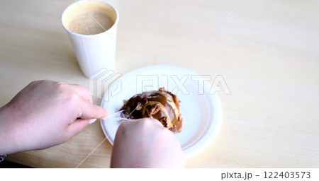 A close-up of a woman's hands slicing a chocolate caramel cinnamon roll disposable cutlery on a white plate coffee in foreground Captures cozy moment perfect showcasing indulgent desserts cafe vibes. 122403573