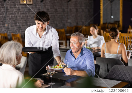 Young male waiter serving dish to elderly couple in restaurant Young male waiter serving dish to elderly couple in restaurant 122408663
