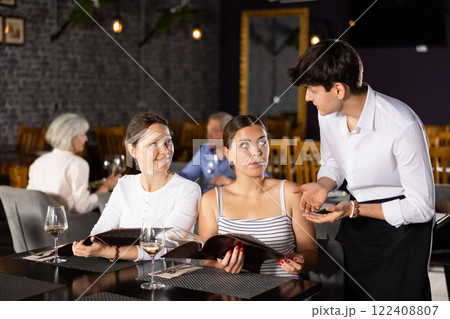 Female guests discussing menu with waiter at table in restaurant 122408807