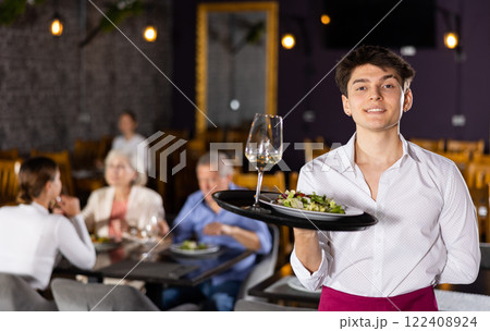 Young waiter guy is standing with order tray in spacious guest room in restaurant. Young waiter guy is standing with order tray in spacious guest room in restaurant. 122408924