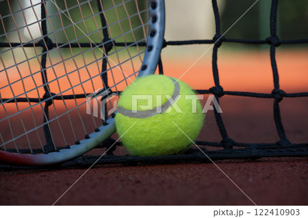 Tennis ball and racket leaning on the net of a clay court Tennis ball and racket leaning on the net of a clay court 122410903