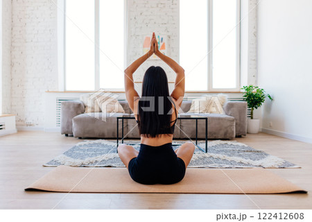 Back view of a woman meditating and doing yoga alone at home in the morning A woman is sitting on a yoga mat in her living room, meditating with her hands raised above her head. 122412608