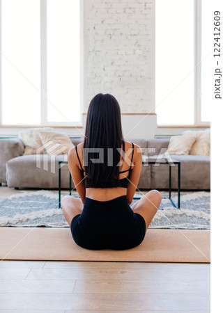 Woman sitting on floor in meditative pose her legs crossed. She is facing away from camera, wearing black workout clothing sports bra and shorts Woman sitting on floor in meditative pose her legs crossed. She is facing away from camera, wearing black workout clothing sports bra and shorts 122412609