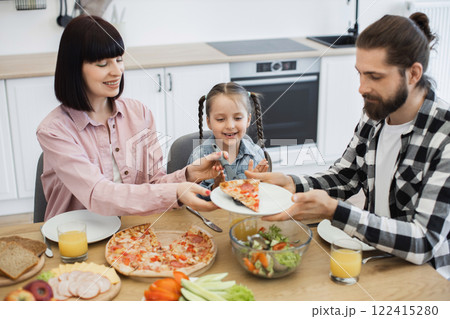 Caucasian family with parents and daughter enjoying pizza breakfast at home. Happy bonding moment around kitchen table with salad and juice. 122415280