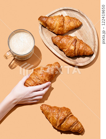 Hand holding fresh croissant on orange background with cappuccino cup and fresh pastry 122419650