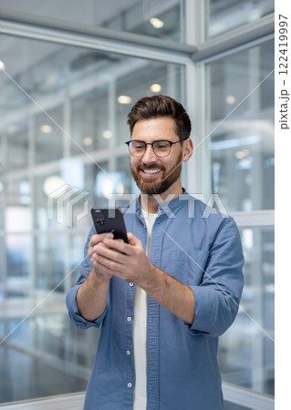 Vertical shot, successful happy man standing inside office using phone. Businessman typing text message and chatting on social media. Vertical shot, successful happy man standing inside office using phone. Businessman typing text message and chatting on social media. 122419997