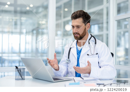 Doctor inside clinic office using laptop for video call, man in white medical coat talking remotely, consulting patients while sitting at desk inside office. Doctor inside clinic office using laptop for video call, man in white medical coat talking remotely, consulting patients while sitting at desk inside office. 122420019