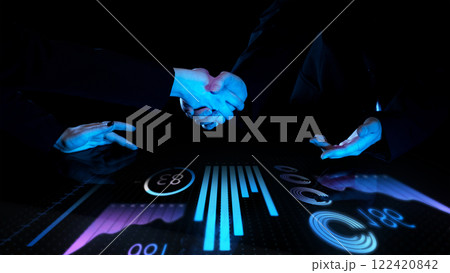 Two professionals shaking hands over futuristic touch screen with digital analytics and bar graphs glowing in blue tones against dark background. 122420842