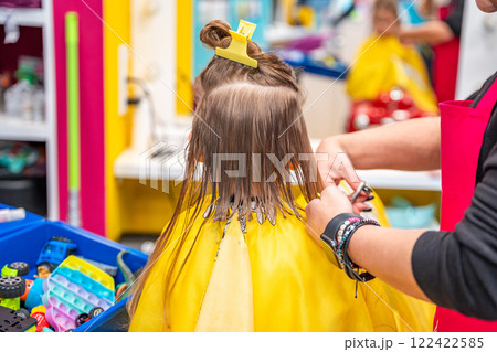 Back view of hairdresser making a hair style to cute little girl in real child salon with colorful interior and toys 122422585