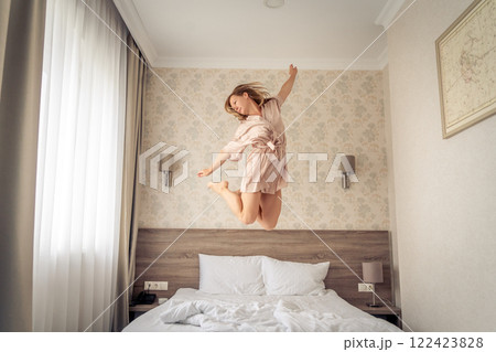Woman having fun and jumping with bathrobe on bed in hotel room 122423828