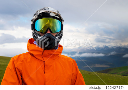 man rides a mountain bike on top of a green hill. A guy in a protective helmet stands on a meadow looking into the distance, with large snow-capped mountains in the background among clouds 122427174