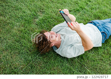 Joyful man smiling and having fun while playing video games with portable console and lying on the grass outdoors. Technology and game addiction concept. Joyful man smiling and having fun while playing video games with portable console and lying on the grass outdoors. Technology and game addiction concept. 122428717