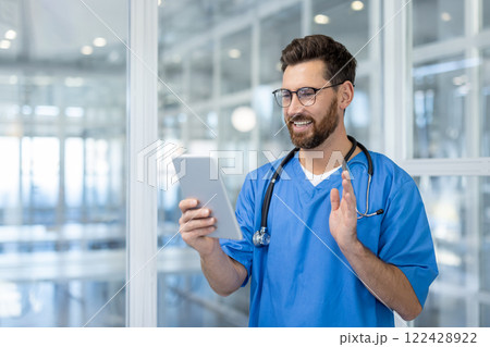 Doctor engaging in video call using tablet, showcasing technology driven healthcare. Medical professional wearing blue scrubs and stethoscope, exuding warmth and confidence in hospital environment. Doctor engaging in video call using tablet, showcasing technology driven healthcare. Medical professional wearing blue scrubs and stethoscope, exuding warmth and confidence in hospital environment. 122428922