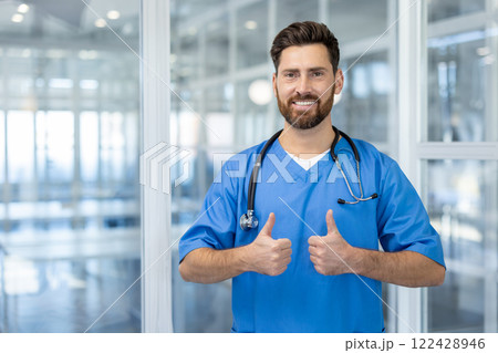 Smiling healthcare professional in blue scrubs giving thumbs up gesture, representing confidence and positivity in medical facility. Stethoscope around neck indicates medical role. 122428946