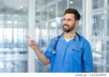 Confident male healthcare professional in blue scrubs with stethoscope indoors. Smiling, pointing doctor embodies medical expertise, positive energy, teamwork in healthcare. Bright, modern setting. Confident male healthcare professional in blue scrubs with stethoscope indoors. Smiling, pointing doctor embodies medical expertise, positive energy, teamwork in healthcare. Bright, modern setting. 122428949