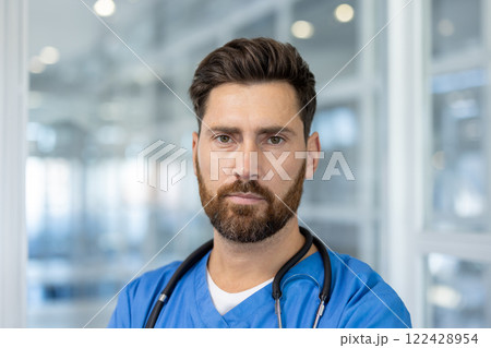 Confident male doctor with beard in blue scrubs and stethoscope in modern hospital setting. Direct eye contact conveys professionalism, dedication, expertise, and trust in medical field. 122428954