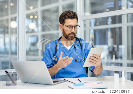 Professional man doctor in blue scrubs interacting with tablet in bright office. Stethoscope around neck indicates healthcare environment. Engaged in virtual consultation or research using technology Professional man doctor in blue scrubs interacting with tablet in bright office. Stethoscope around neck indicates healthcare environment. Engaged in virtual consultation or research using technology 122428957