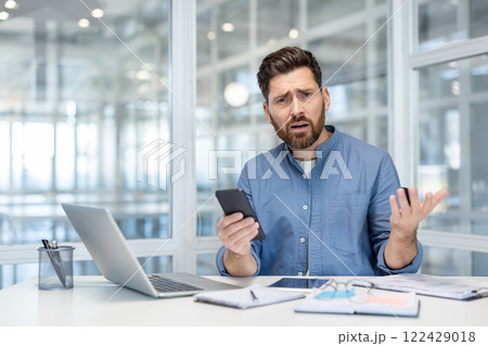 Upset and disappointed man with phone in hands. Office worker at workplace looking anxiously at camera, working with laptop inside office at workplace. Upset and disappointed man with phone in hands. Office worker at workplace looking anxiously at camera, working with laptop inside office at workplace. 122429018