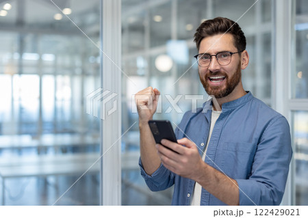 Portrait of a successful businessman at workplace inside office, a man with a phone in his hands celebrates victory and successful results of achievement, holds his hand up and looks at the camera. Portrait of a successful businessman at workplace inside office, a man with a phone in his hands celebrates victory and successful results of achievement, holds his hand up and looks at the camera. 122429021