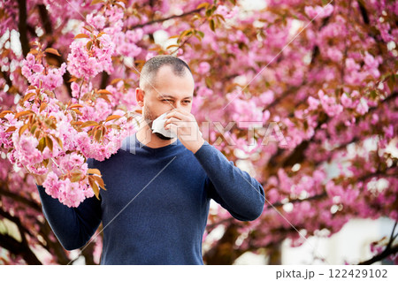 Man allergic suffering from seasonal allergy at spring in blossoming garden at springtime. Man sneezing and blowing nose using nasal handkerchief in front of blooming tree. Spring allergy concept. Man allergic suffering from seasonal allergy at spring in blossoming garden at springtime. Man sneezing and blowing nose using nasal handkerchief in front of blooming tree. Spring allergy concept. 122429102