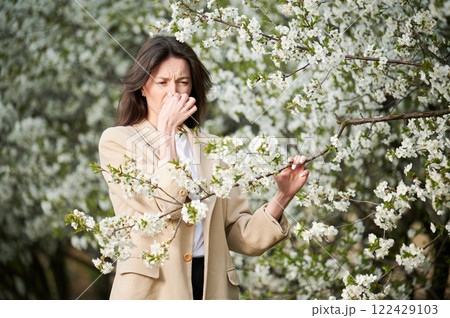 Woman allergic suffering from seasonal allergy at spring in blossoming garden at springtime. Young woman sneezing, having runny nose in front of blooming tree. Spring allergy concept Woman allergic suffering from seasonal allergy at spring in blossoming garden at springtime. Young woman sneezing, having runny nose in front of blooming tree. Spring allergy concept 122429103