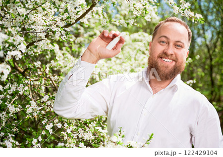 Man allergic using medical nasal drops, suffering from seasonal allergy at spring in blossoming garden. Handsome smiling man showing medicament near blooming tree outdoors. Spring allergy concept. 122429104