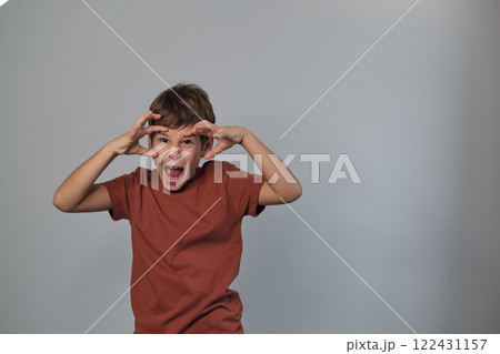A boy in a rust-colored shirt makes a monster face, fingers pulling his eyes wide. This playful moment reflects childhood imagination and the joy of pretend play. A boy in a rust-colored shirt makes a monster face, fingers pulling his eyes wide. This playful moment reflects childhood imagination and the joy of pretend play. 122431157