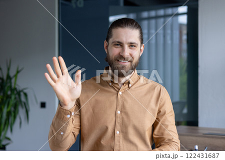 Friendly man wearing a brown shirt waves towards the camera, expressing positivity and approachability, situated in a modern office environment. Friendly man wearing a brown shirt waves towards the camera, expressing positivity and approachability, situated in a modern office environment. 122431687