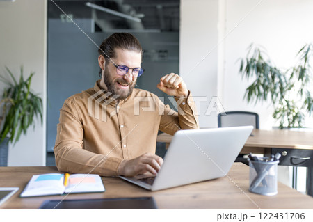 A young man with glasses and a beard expressing excitement working on a laptop, showing a celebratory fist. Perfect for depicting success, achievement, and positivity in a professional workspace. A young man with glasses and a beard expressing excitement working on a laptop, showing a celebratory fist. Perfect for depicting success, achievement, and positivity in a professional workspace. 122431706
