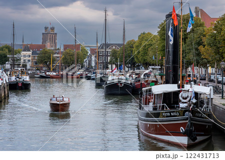 Dordrecht, South Holland, Holland - 09.07.2024: Serene harbor with moored boats under a dramatic cloudy sky. Quaint historic buildings and trees line the waterfront, reflecting in the calm green water 122431713