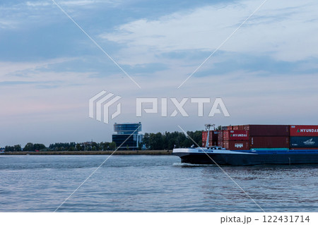Dordrecht, South Holland, Holland - 09.07.2024: A cargo ship named "Devotion" transports Hyundai shipping containers along a calm river. A modern glass office building stands on the shore, with trees Dordrecht, South Holland, Holland - 09.07.2024: A cargo ship named "Devotion" transports Hyundai shipping containers along a calm river. A modern glass office building stands on the shore, with trees 122431714