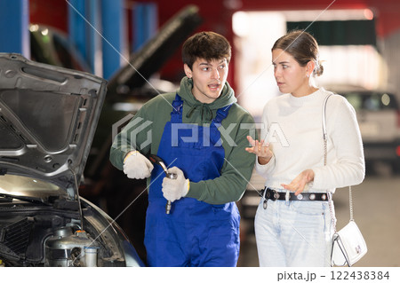 Mechanic advises young woman client on repairing under hood of car in car service station Mechanic advises young woman client on repairing under hood of car in car service station 122438384