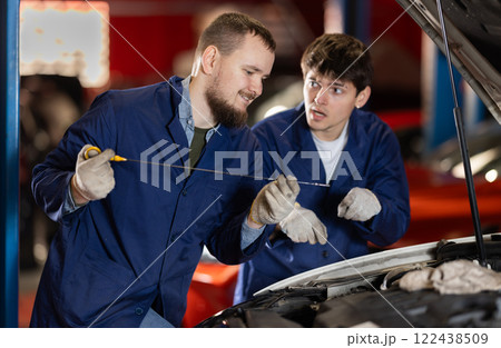 Two auto mechanics checking engine oil level under the hood of car together in garage Two auto mechanics checking engine oil level under the hood of car together in garage 122438509