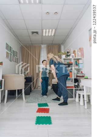 boy smiling and looking at therapist during a sensory motor exercise in a bright therapy room with balance mats on the floor, concept of childhood development, physiotherapy boy smiling and looking at therapist during a sensory motor exercise in a bright therapy room with balance mats on the floor, concept of childhood development, physiotherapy 122439537