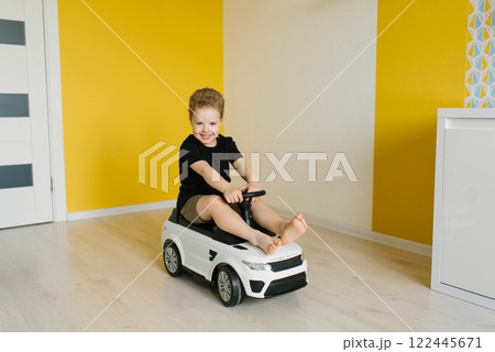 Child boy is sitting in toy car, smiling and enjoying himself in room is brightly lit and has yellow wall, creating cheerful atmosphere Child boy is sitting in toy car, smiling and enjoying himself in room is brightly lit and has yellow wall, creating cheerful atmosphere 122445671