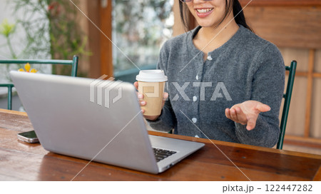 A smiling Asian woman discusses work through an online video meeting, working remotely from a cafe. A smiling Asian woman discusses work through an online video meeting, working remotely from a cafe. 122447282