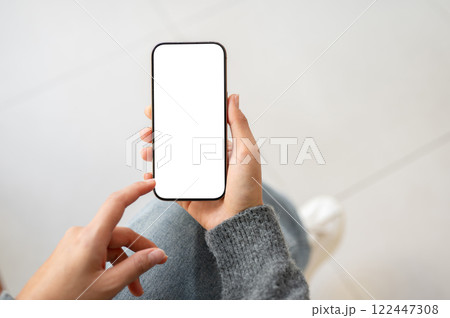 A top view of a woman holds a smartphone with a white screen mockup while sitting indoors. 122447308