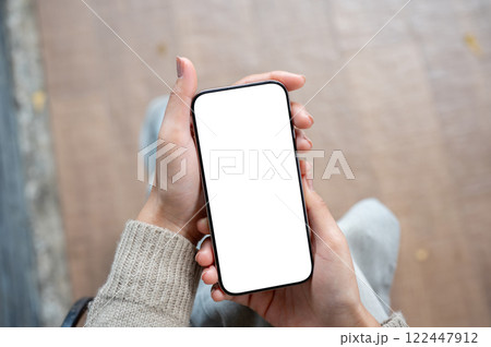A top view of a woman sits indoors and holds a smartphone with a white screen mockup. 122447912