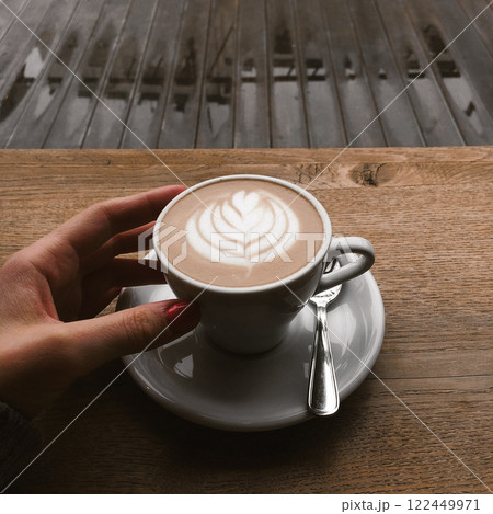 White coffee cup stands on wooden table on cafe terrace on a city street. Morning coffee outdoors in rainy day. Cappuccino, latte cup in female hand. 122449971