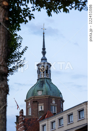 A historic clock tower with a green dome and spire rises above brick and modern buildings. The evening sky and tree branches frame the scene, adding depth to the European architectural setting. A historic clock tower with a green dome and spire rises above brick and modern buildings. The evening sky and tree branches frame the scene, adding depth to the European architectural setting. 122451086