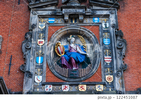 A historic architectural relief on a red brick wall features a statue of a seated woman in blue robes holding a shield and a golden palm leaf, surrounded by ornate stonework and heraldic crests. A historic architectural relief on a red brick wall features a statue of a seated woman in blue robes holding a shield and a golden palm leaf, surrounded by ornate stonework and heraldic crests. 122451087