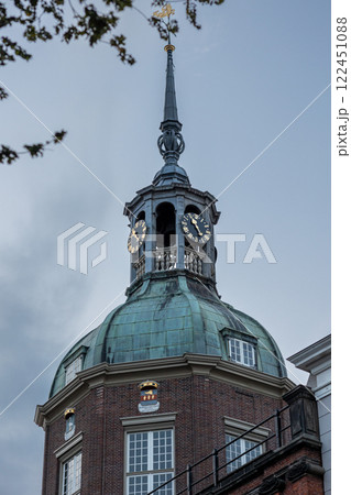 A historic clock tower with a green dome and spire rises above brick and modern buildings. The evening sky and tree branches frame the scene, adding depth to the European architectural setting. A historic clock tower with a green dome and spire rises above brick and modern buildings. The evening sky and tree branches frame the scene, adding depth to the European architectural setting. 122451088
