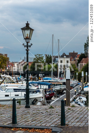A vintage street lamp stands in the foreground near a harbor filled with boats. The scene captures a European coastal town with historic buildings, docks, and calm waters under an overcast sky. 122451089
