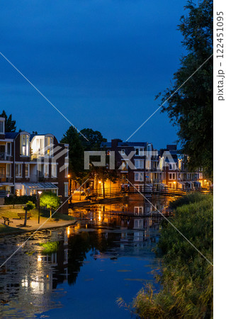 A serene night scene of a modern European neighborhood with illuminated buildings reflecting in the calm canal water. A small bridge connects the houses, while streetlights contrast with the blue sky. 122451095